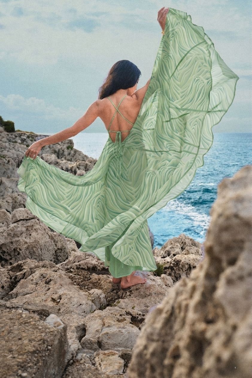 Woman in flowing green dress standing on rocky coastline overlooking the ocean